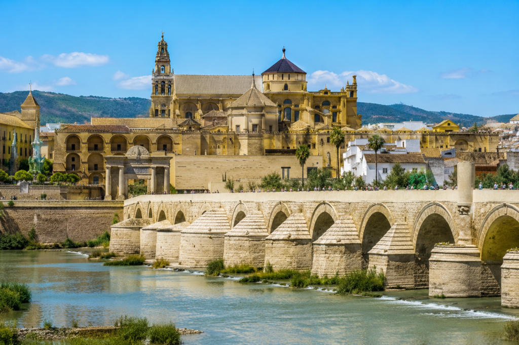 The Roman Bridge and the Mosque-Cathedral (Mezquita) in Córdoba, Spain, viewed across the Guadalquivir River on a sunny day.